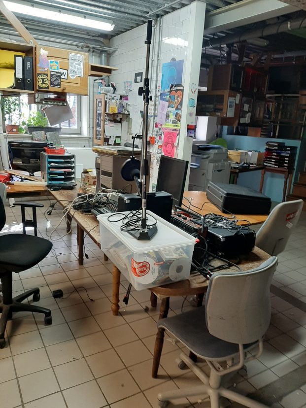 Colour photograph of the original spot take for the nascent Radio WORM, taken in the old downstairs office at WORM in summer 2020. A photograph of tables, chairs, cupboards and radio equipment. WORM's office is its usual cramped and cluttered state.