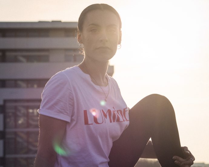 Photograph of Myriam Meret, with a shining sun and a tower block behind her. Used to promote the WORM Pirate Bay residency focussing on rest, led by Myriam Meret. October 2020