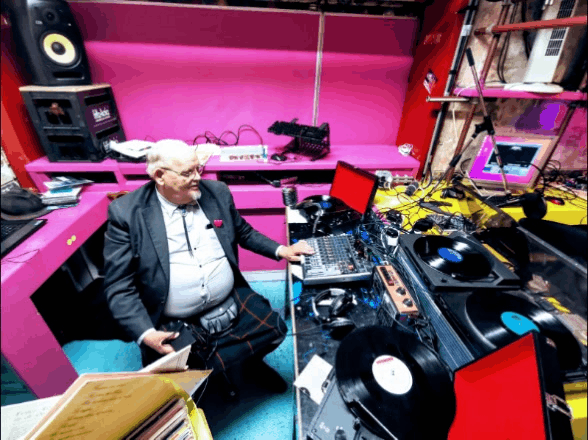 Colour photograph of an older person in a grey suit, shirt and kilt and Slim Jim necktie, sitting in a predominantly pink space, surrounded by recording and broadcasting equipment.