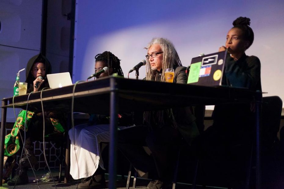 Colour photograph of three Black women and one Black man sitting on a stage at a table with microphones in WORM Central Station with a large screen behind them. Image commemorating WORM's Afrofuturism Now! Festival November 2015.