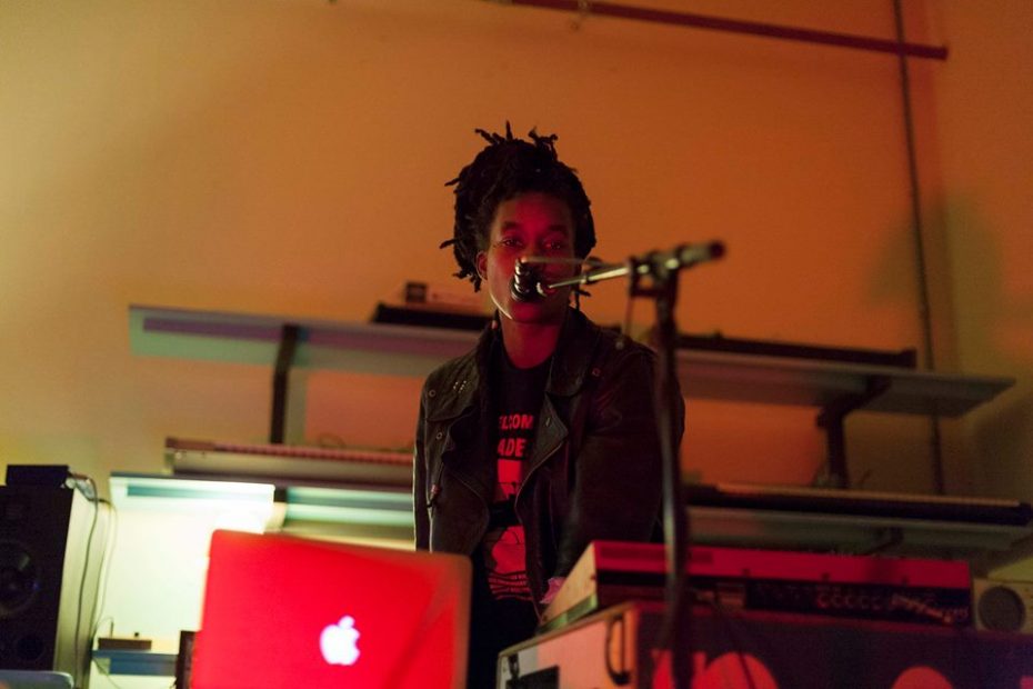 Colour photograph of a Black woman standing behind a laptop and shouting into a stand microphone on a stage, contrasted against a red-lit background in maybe WORM Sound Studios. Image commemorating WORM's Afrofuturism Now! Festival November 2015.