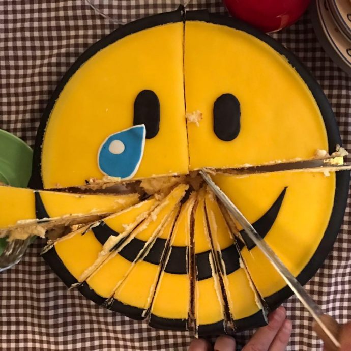 A picture of a round cake, styled as a classic smiley (yellow face, black eyes and myth) with a blue tear rolling down one cheek. The cake has been cut into slices along its bottom half but no slice has been removed. Taken to commemorate the leaving of Creative Director Hajo Doorn in 2017.