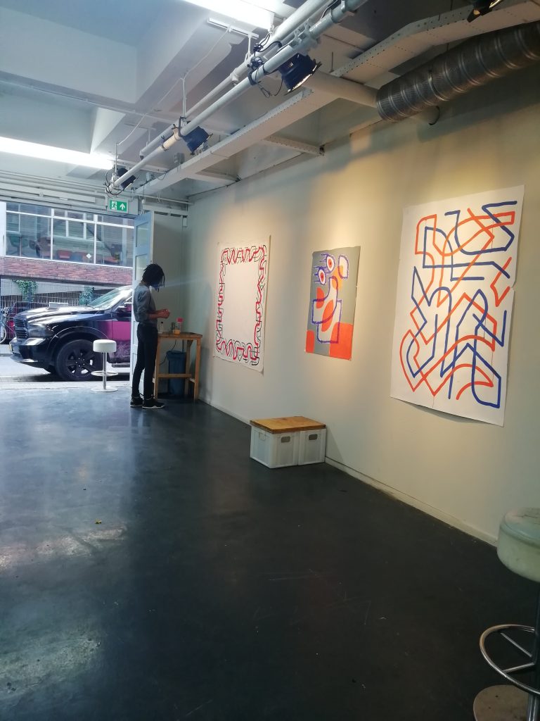 A colour photograph of the Slash Gallery with the doors fully open. To the right of the image, three large colour drawings can be seen. A figure is near the door in the corner of the room, looking at some text. Part of Iloy Drisdale's residency, summer 2020.