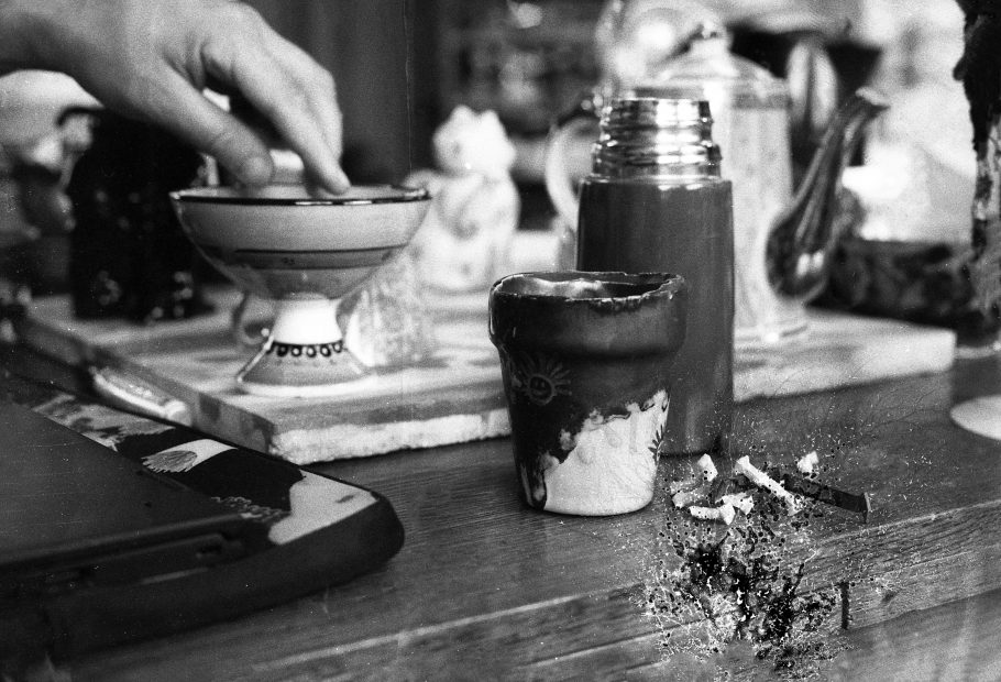 Black and white photograph of a hand hovering over a bowl, with diverse array of kitsch ceramics out of focus n the background. An unscrewed flask and a cup are in the foreground. Used in a WORM interview with the artist Susanne Khalil Yussef 2018.