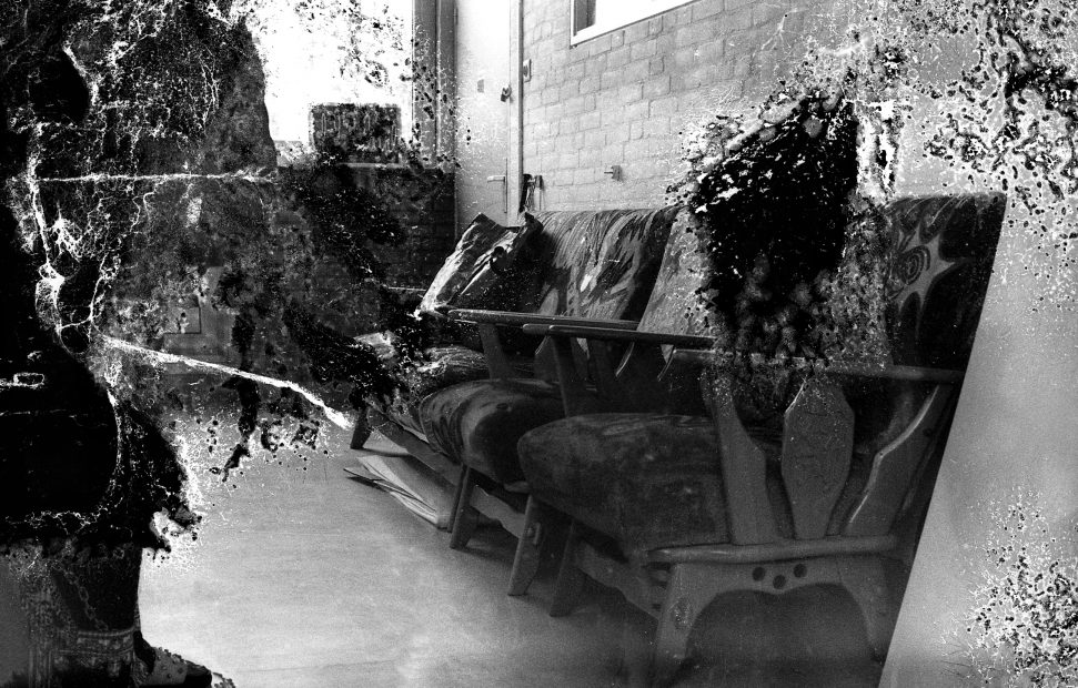 A treated black and white photograph, maybe deliberately treated to suggest a damaged print of a brick walled interior. Sturdy wooden chairs line the wall. Used in a WORM interview with the artist Susanne Khalil Yussef 2019.
