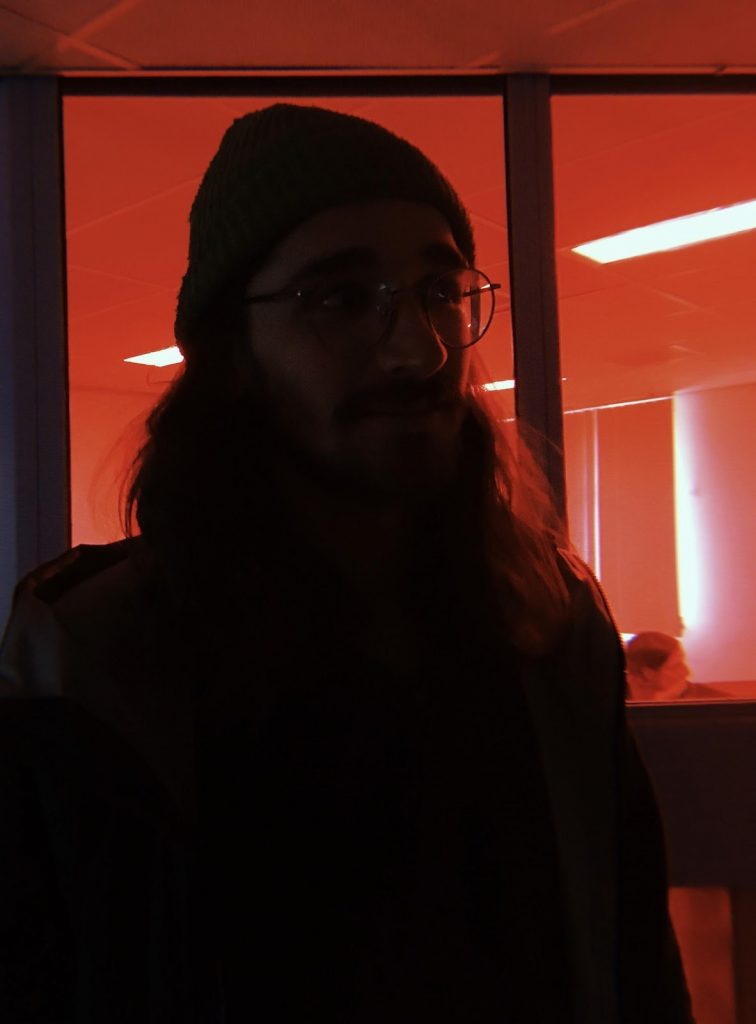 A colour photograph of a bearded and bespectacled person wearing a woollen hat, looking at the camera, their features in shadow and a strong red light behind them. Portrait of Jacques Kayser, the organiser of the Sonaural Festival March 2020.