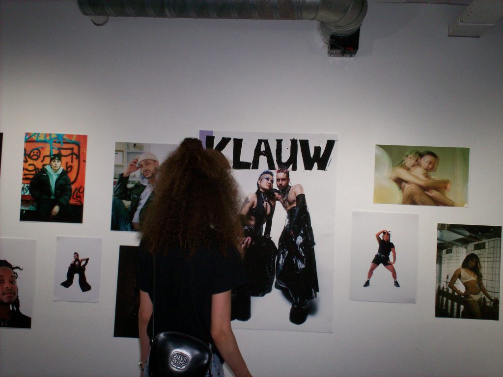 A photograph of a person with long frizzy hair and back to the camera, looking at various photographs on a white wall. One photograph says KLAUW at the top of it.