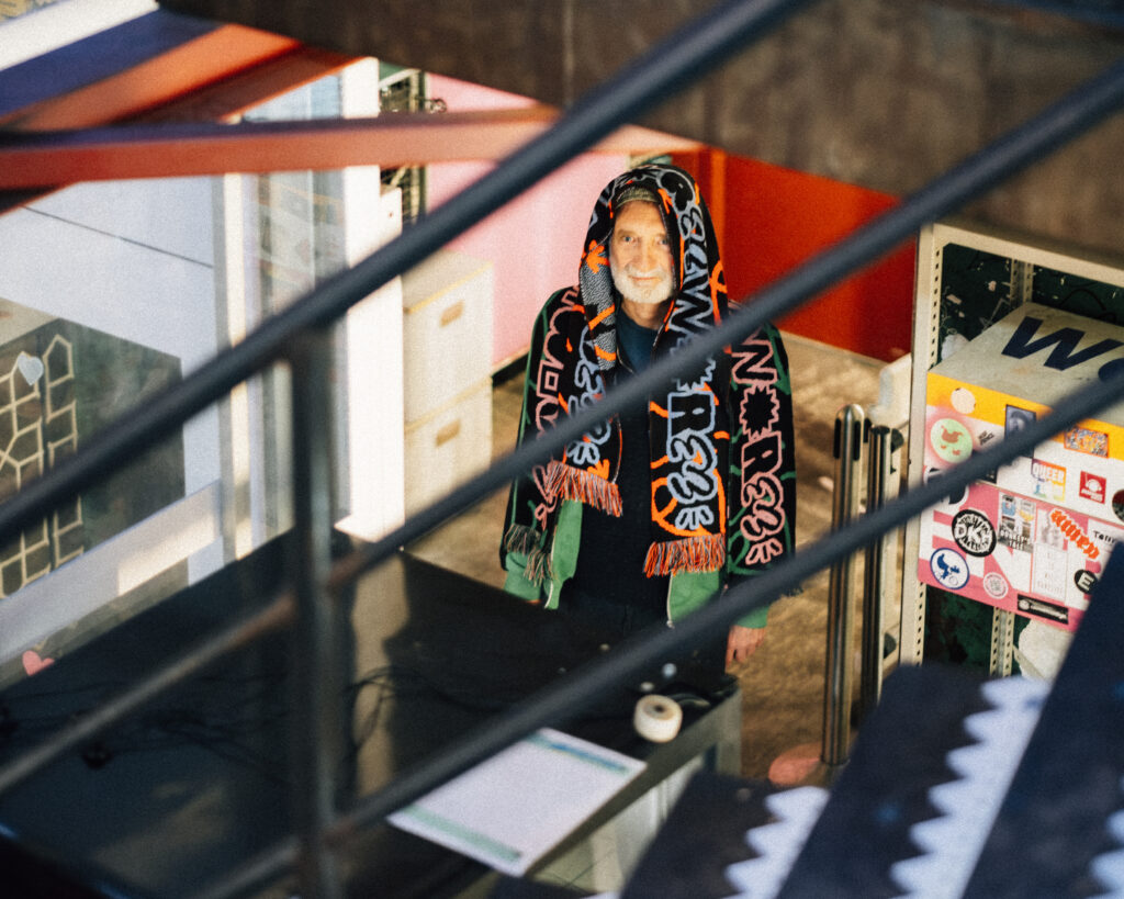 A colour photograph - taken from an angle looking down from some stairs - of one standing white-bearded man, his head fully draped in WORM scarves.