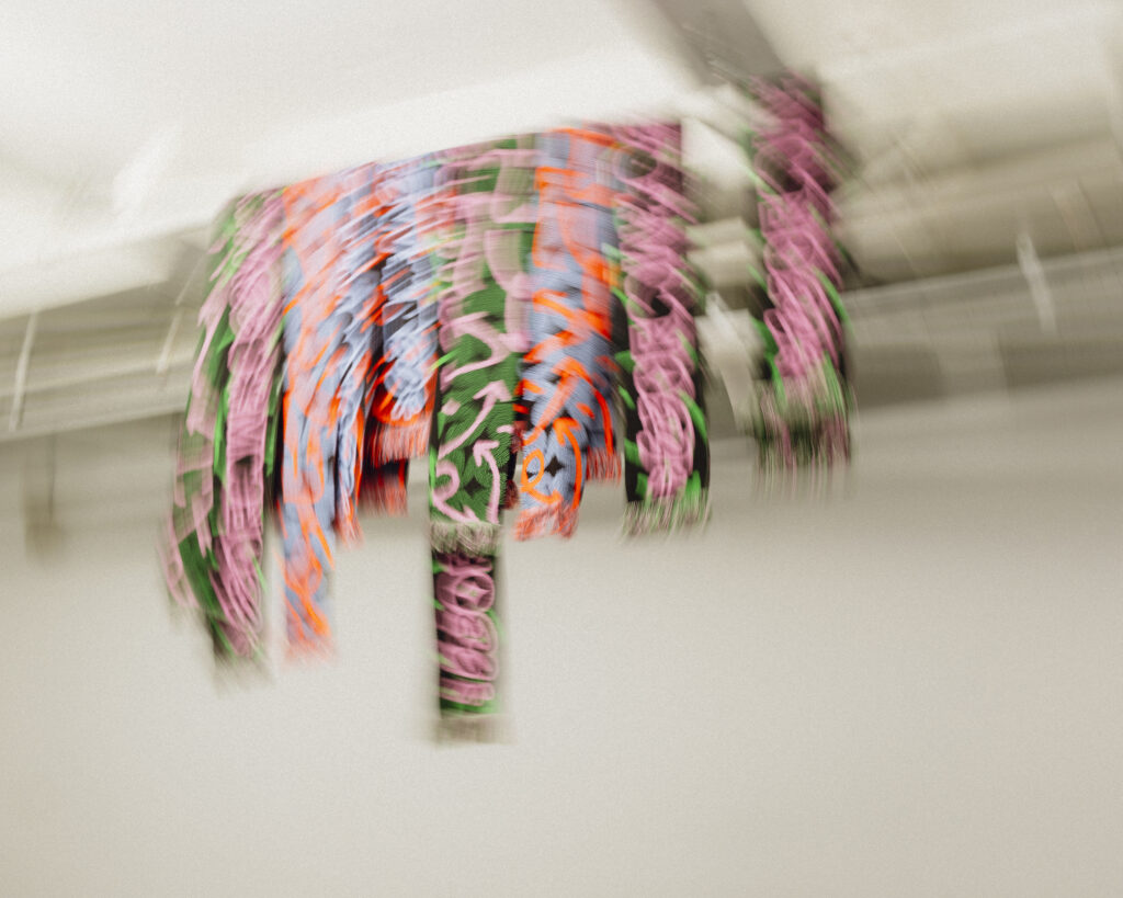 Blurred colour photograph - with a white background of all the WORM scarves hanging on a support