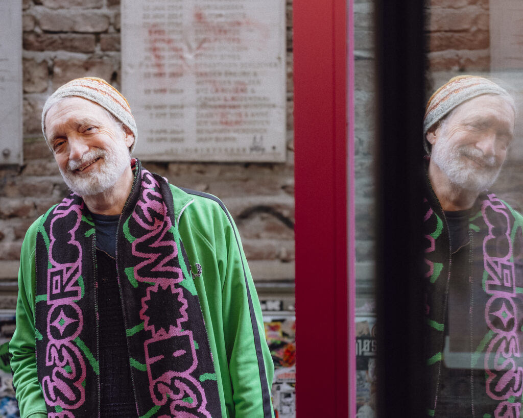 A colour photograph - taken outside WORM - of a standing white-bearded man wearing a woollen hat and a green tracksuit top - with a pink and black WORM scarf around his shoulders.