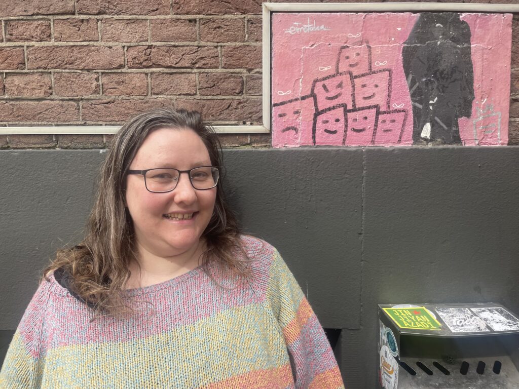 Colour portrait photograph of Art Gang's Mimi van Rinsum standing outside WORM (next to the grey-painted wall) smiling.