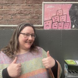 Colour portrait photograph of Art Gang's Mimi van Rinsum standing outside WORM (next to the grey-painted wall) smiling and with her thumbs up.