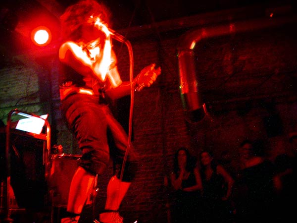 Colour photo of a man wearing a wig singing, knees bent, in a radium glow of light onstage. Part of the WORM tribute to Wim T. Schippers.