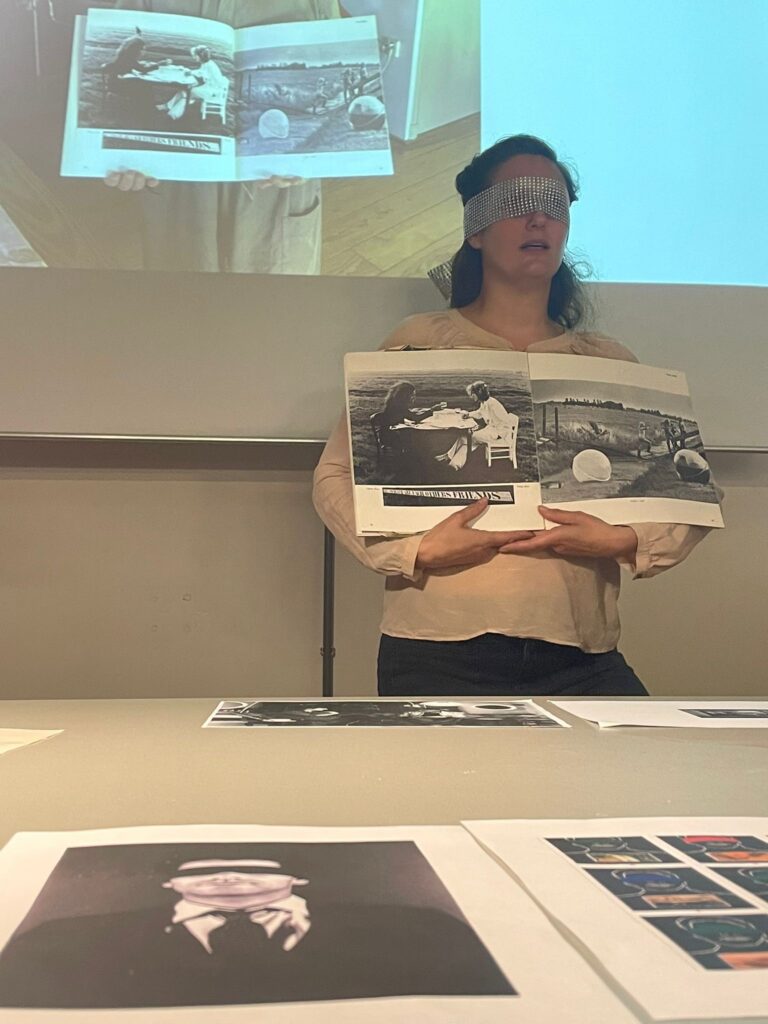 Nina Boas blindfolded, presenting her ongoing research project The Improvised Family. In her hands is the book Lucht, Water, Vuur, Aarde (Air, Water, Fire, Earth) by Fons Elders, shown enlarged in the projection behind her. The book features artists Flavio Pons and Philip Boas (her father) and was published in 1977. In the foreground is an image of Claudio Goulart.