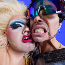 Drek Manor collective with blue background. Two faces on the foreground. One with blonde hair and heavy blue eye makeup and red lips and one with ski big glasses showing their theeth.