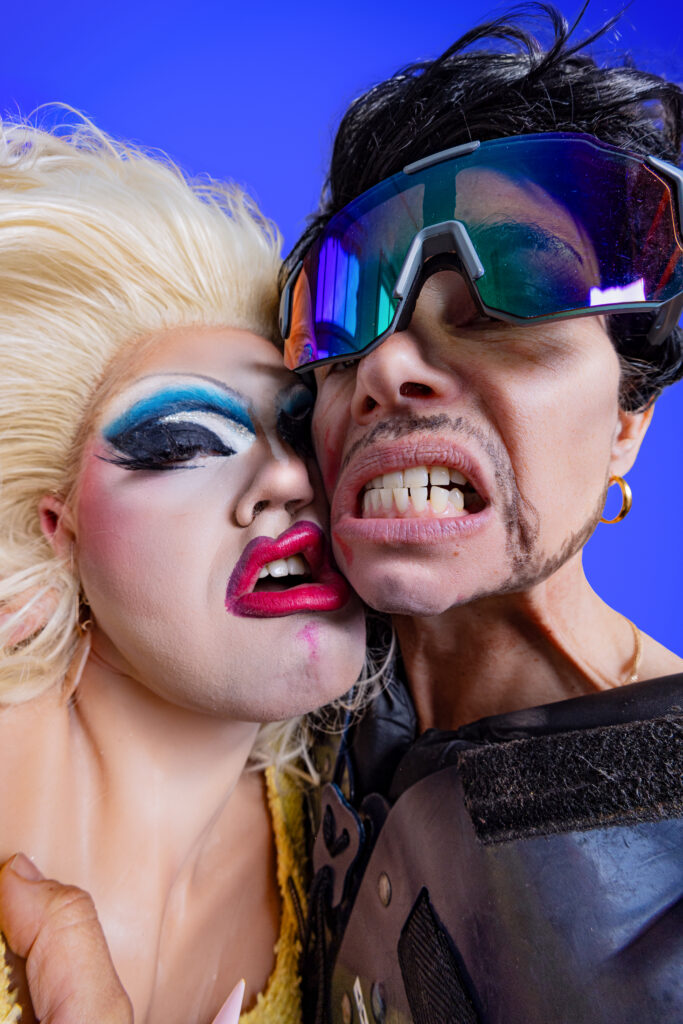 Drek Manor collective with blue background. Two faces on the foreground. One with blonde hair and heavy blue eye makeup and red lips and one with ski big glasses showing their theeth.