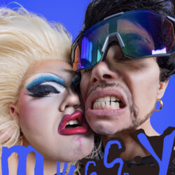 Drek Manor collective with blue background. Two faces on the foreground. One with blonde hair and heavy blue eye makeup and red lips and one with ski big glasses showing their teeth.